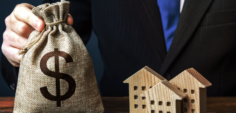 businessman holding burlap money bag next to wooden residential figurines symbolic of long term loan