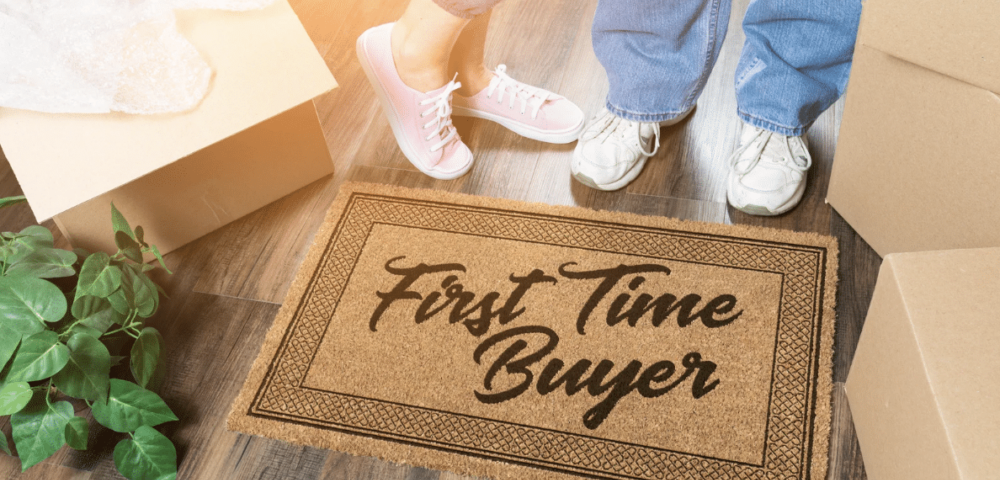 first-time home buyers concept. couple's shoes in front of welcome mat that says "first time buyer" surrounded by moving boxes