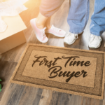 first-time home buyers concept. couple's shoes in front of welcome mat that says "first time buyer" surrounded by moving boxes