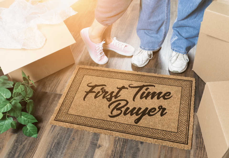 first-time home buyers concept. couple's shoes in front of welcome mat that says "first time buyer" surrounded by moving boxes